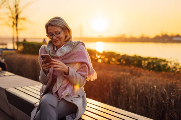 istock Senior adult woman using smartphone while sitting on park bench, overlooking river 2174384415