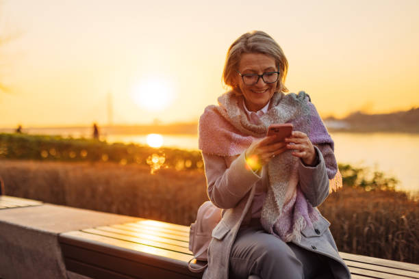 istock Carefree senior woman using mobile phone while sitting on park bench, overlooking river 2174384429
