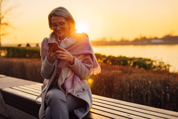 istock Smiling senior woman using mobile phone while sitting on park bench, overlooking river 2174384475