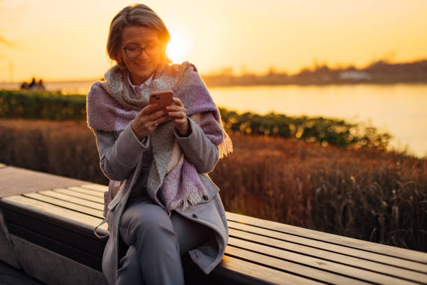 istock Cheerful senior woman using mobile phone while sitting on park bench, overlooking river 2174384500