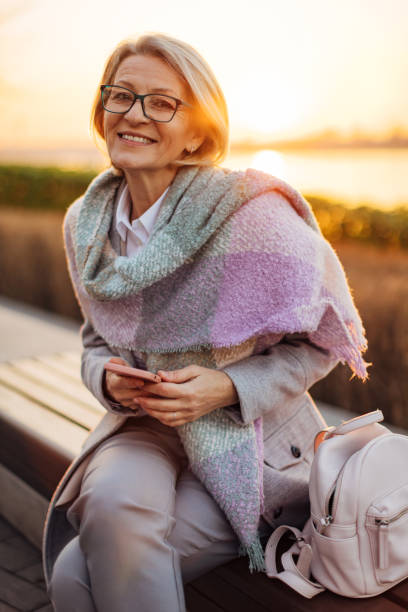 istock Smiling senior woman looking directly at the camera while sitting on a park bench overlooking the river during sunset 2174384594