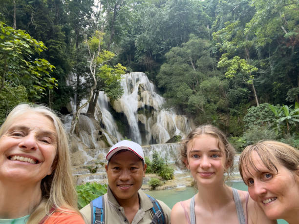 A selfie taken on a mobile phone of a mother and daughter with their tour guide and family friend taking a picture at a local beauty spot in Luang Prabang in Laos in southeast Asia the waterfall they are visiting is called Kuang Si Waterfall. They are admiring the beauty and strength in nature and taking selfies for their memories.