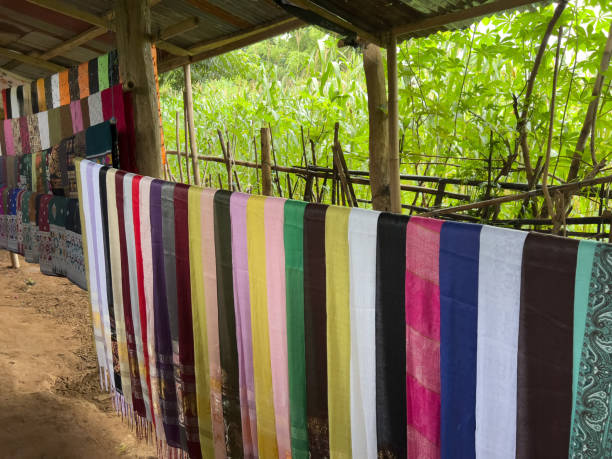 Medium shot of a selection of multicoloured handmade silk scarves displayed inside a wooden shelter, available for tourists to purchase whilst visiting the area near Luang Prabang, Laos. Behind the shelter there is an abundance of lush green foliage creating a screen.