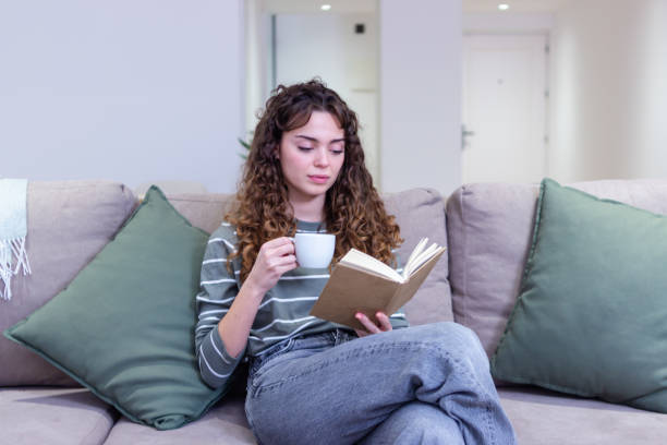 istock Young woman is reading a book and drinking coffee on the sofa at home 2174406238