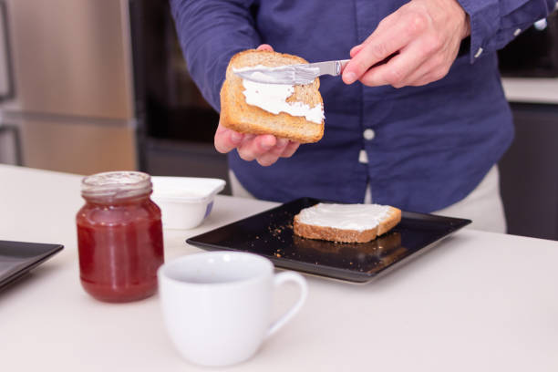 Man is spreading cream cheese on toast, preparing a quick breakfast in his kitchen