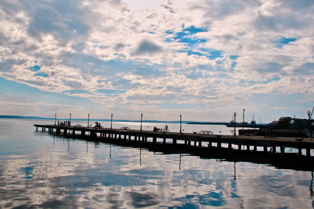The image features a serene pier extending over calm waters, reflecting the partly cloudy sky above. The pier is lined with vintage-style lampposts and is occupied by a few people engaging in various leisurely activities, such as walking and biking. The still water beautifully mirrors the sky, creating a sense of tranquility. In the distance, the horizon stretches across the water, with hints of industrial structures or distant ships, possibly indicating a harbor area. The overall atmosphere is peaceful, with soft lighting from the overcast sky enhancing the calmness of the scene.