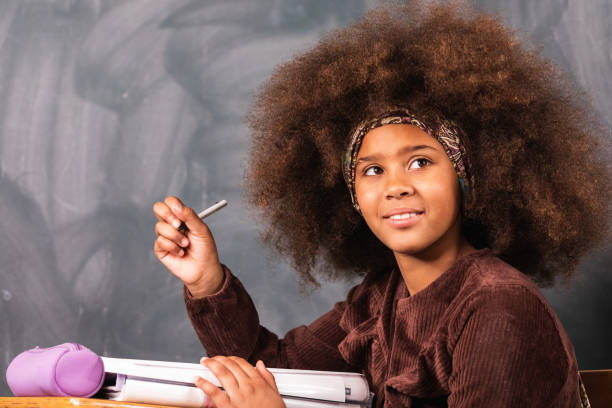 Side view of clever teenage African American girl sitting at table in classroom with blackboard during lesson at school