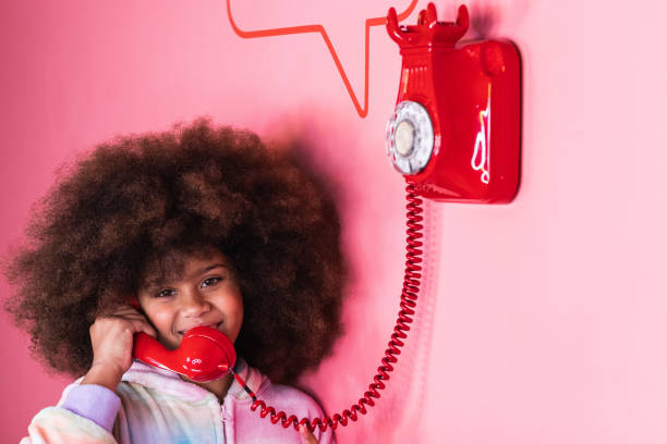 Smiling ethnic girl with Afro hairstyle talking on old fashioned red telephone in studio and looking at camera on pink background