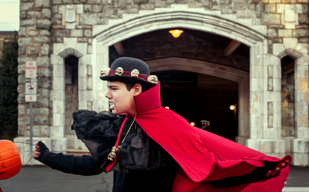 Young boy wearing black outfit with bright red cape and decorative hat runs excitedly towards stone castle entrance. Arched doorway and stone walls create mysterious atmosphere, complemented by orange pumpkin decoration in foreground. Halloween theme evident in costume and setting.