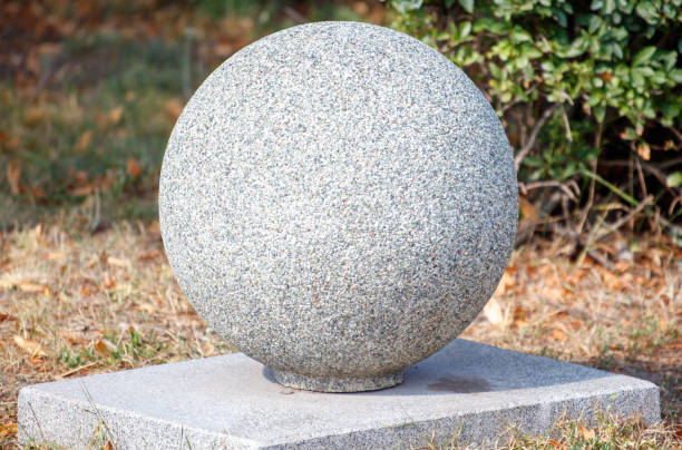 A large grey stone ball sits on a concrete base. The stone ball is surrounded by a patch of grass and some bushes. The scene has a calm and peaceful atmosphere, with the stone ball as the focal point
