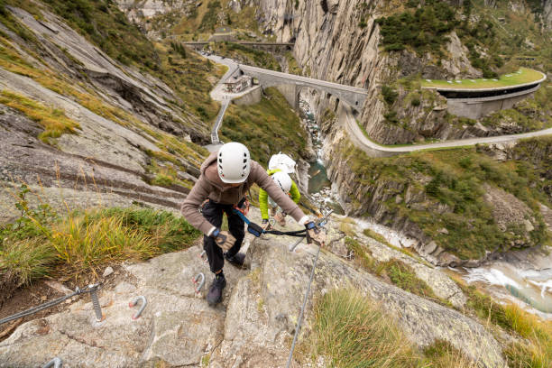 Two climbers wearing helmets and harnesses ascending a steep mountain trail with safety cables, surrounded by scenic mountain roads and a river gorge.