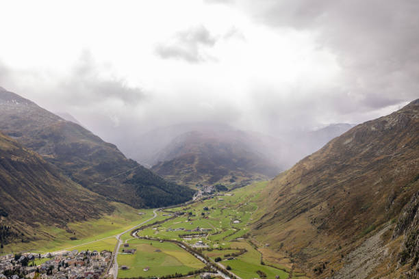 Scenic view of a misty valley in Switzerland, framed by dramatic clouds and rolling hills, with a small village nestled below.