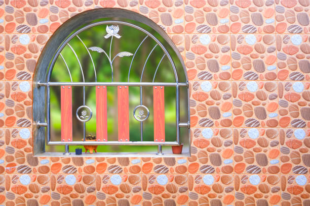 Beige tile fence wall background with pebble stone pattern and decorative stainless steel wrought iron inside of arch window in front of home garden of vintage house
