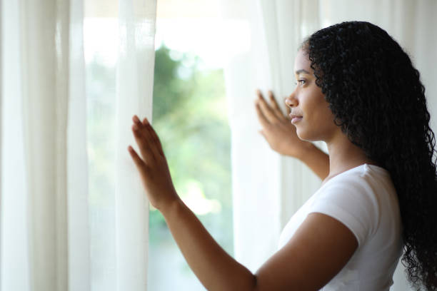 Serious black woman opening curtains at home