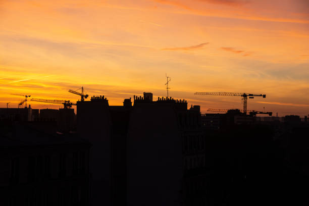 This image captures the serene beauty of an urban skyline at sunset. The sky is painted with hues of orange, yellow, and soft pink as the sun dips below the horizon. Silhouettes of buildings, characterized by traditional European architecture, stand in stark contrast against the vibrant sky. In the distance, cranes tower above the city, hinting at ongoing development and the ever-changing nature of the urban landscape. The scene evokes a sense of calm, blending the tranquility of nature with the bustling energy of the city.