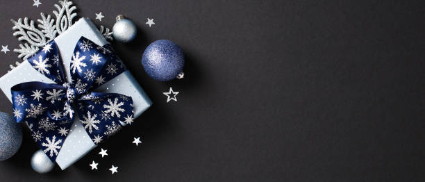 istock Top view of Christmas gift box with blue snowflake-patterned ribbon bow., blue and silver Christmas baubles, snowflakes, confetti on black background. 2174611785