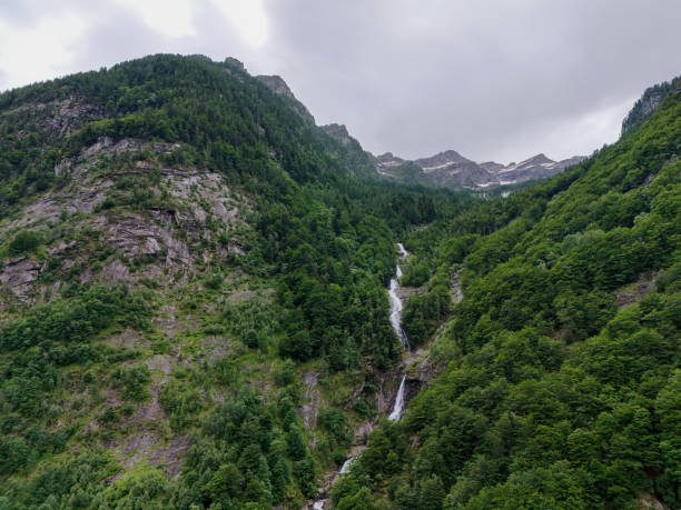 A tall waterfall flows through a densely forested mountain, surrounded by lush greenery, with the rocky peaks visible in the background under a cloudy sky.