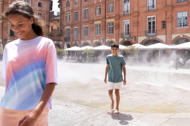 An over-the-shoulder shot of a young boy following his sister out of a water mist fountain. They are cooling down in a lively, open square with arched brick buildings in the background. They are enjoying their holiday together in Montauban near Toulouse, France. \n\nVideos are available similar to this scenario.