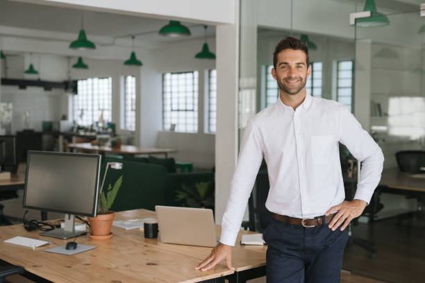 istock Smiling young businessman leaning against a table in an office 2174636846