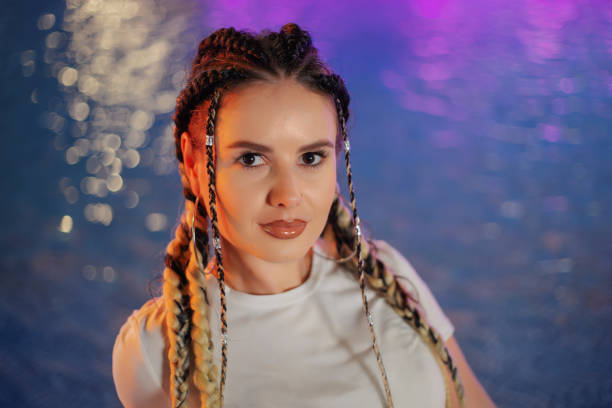 A young woman with stylish pigtails confidently poses at night by the illuminated water, demonstrating modern fashion and beauty.