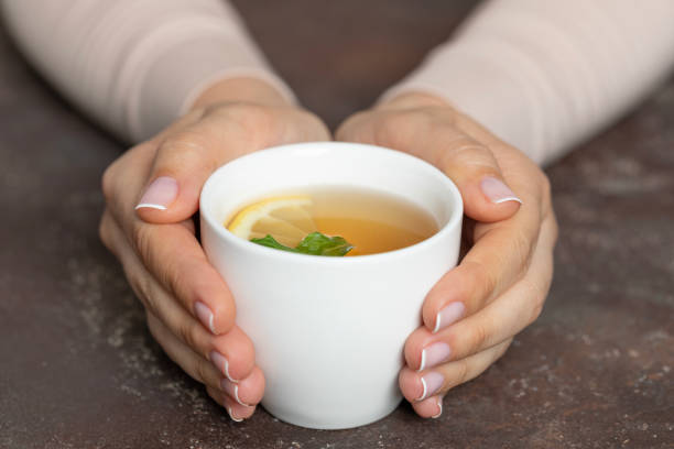 A close-up shot of a person's hands gently holding a white cup filled with herbal tea, garnished with a slice of lemon and fresh mint leaves. The scene suggests warmth, relaxation, and a moment of calm, perfect for promoting wellness and mindfulness. Ideal for health, wellness, and self-care content.