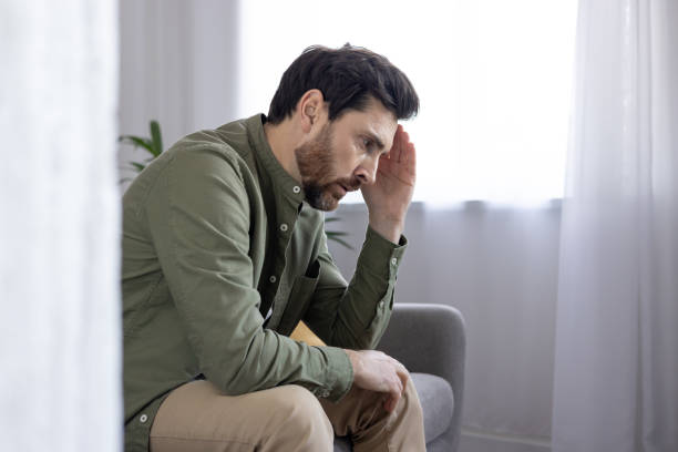 istock Stressed man sitting on a couch holding his head and looking worried in a bright room 2174659582