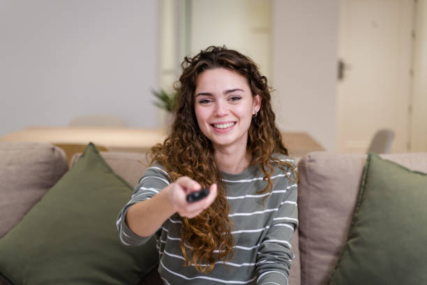 istock Young woman sitting on couch watching tv and holding remote control 2174659868