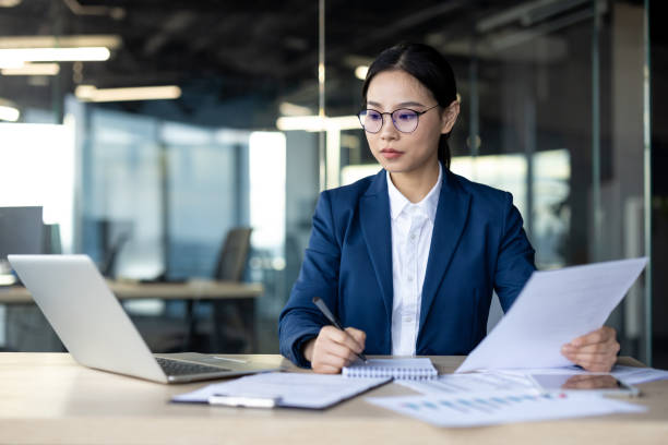 istock Serious businesswoman analyzes financial documents in modern office setting, focusing on paperwork while using laptop and taking notes. 2174663317