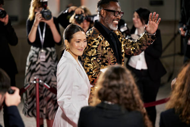Man in tuxedo arm in arm with woman wearing long gown, smiling and interacting during interview, photographers in background.