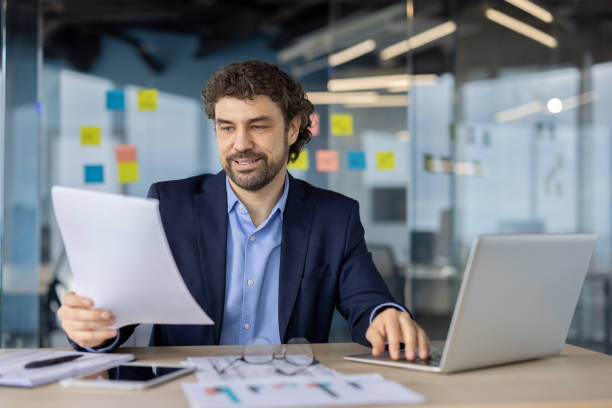 istock Confident businessman in modern office reviewing documents and working on laptop, showcasing productivity and focus in professional setting with technology and innovation 2174666522