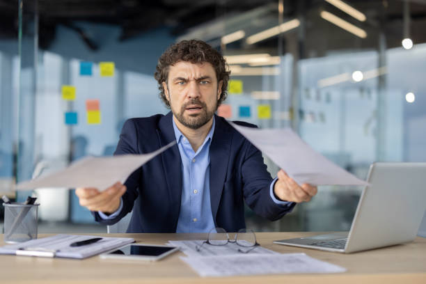istock Businessman stressed with papers in modern office setting, laptop and smartphone visible on desk 2174667552