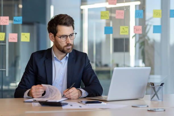 istock Serious financier working in modern office reviewing documents and analyzing data at laptop with phone nearby 2174670002