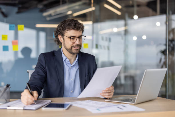 istock Smiling businessman working at office desk reviewing documents with laptop and tablet by side 2174671475