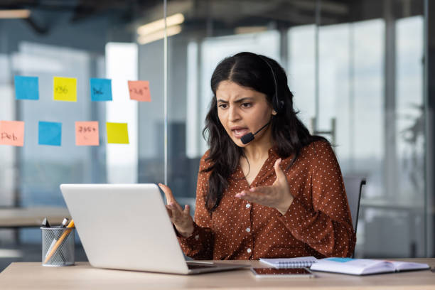 Angry woman yelling on online video call, support worker with headset working inside office using laptop, frustrated with interlocutor