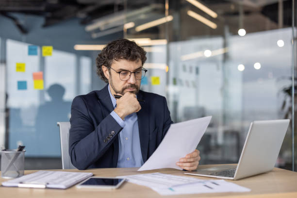 istock Businessman analyzing documents in modern office with laptop and smartphone on desk, contemplating financial data 2174676770