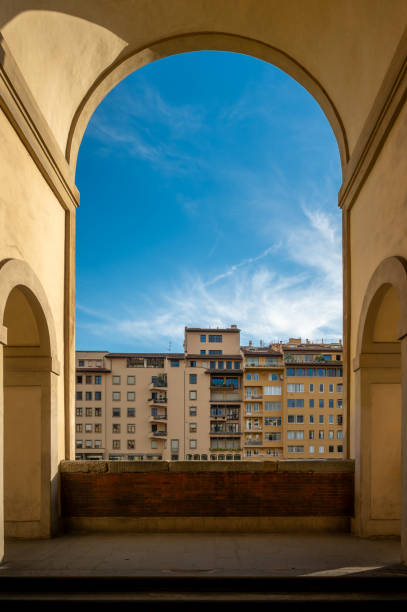 A picturesque view of residential buildings through a historical arched corridor near Ponte Vecchio bridge in Florence, Italy. The sunlight enhances the architectural details, and the blue sky provides a stunning backdrop, capturing the essence of Italian urban beauty and classical design.