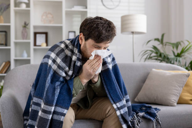 istock Man feeling sick at home wrapped in blanket sneezing into tissue, capturing concepts of illness, cold, flu, and comfort in home environment 2174678586