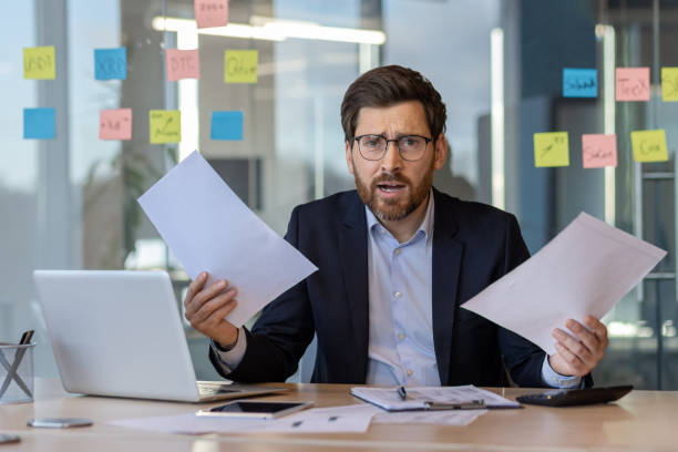 istock Confused businessman holding paperwork in modern office with computer, analyzing data on phone, working on project 2174681461