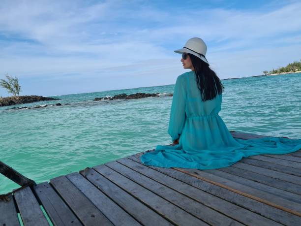A serene moment by the water, where a woman sits peacefully on a dock, wearing a flowy blue dress and a white hat, enjoying the coastal view and gentle waves.