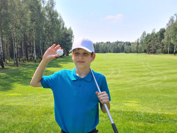 On a sunny afternoon, a young boy stands on a well-maintained golf course. He confidently holds a golf ball in one hand, ready to make his next move as he enjoys the game.