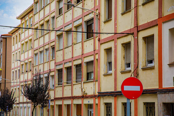 The image showcases a multi-story residential building with a classic, rustic appearance characterized by alternating brick and white concrete walls. The structure is typical of an urban or suburban neighborhood, with multiple windows evenly spaced across the façade. Several balconies are adorned with clotheslines where colorful laundry hangs to dry, adding a vibrant and lived-in feel to the scene. The building's exterior shows signs of age, with some worn areas and peeling paint, reflecting its long-standing presence in the area. There are small details such as electrical wires, street lamps, and the tops of iron fences visible in the foreground. The overall composition captures the essence of everyday life in a residential neighborhood, highlighting the charm and authenticity of community living in an older apartment complex.