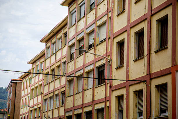 The image showcases a multi-story residential building with a classic, rustic appearance characterized by alternating brick and white concrete walls. The structure is typical of an urban or suburban neighborhood, with multiple windows evenly spaced across the façade. Several balconies are adorned with clotheslines where colorful laundry hangs to dry, adding a vibrant and lived-in feel to the scene. The building's exterior shows signs of age, with some worn areas and peeling paint, reflecting its long-standing presence in the area. There are small details such as electrical wires, street lamps, and the tops of iron fences visible in the foreground. The overall composition captures the essence of everyday life in a residential neighborhood, highlighting the charm and authenticity of community living in an older apartment complex.