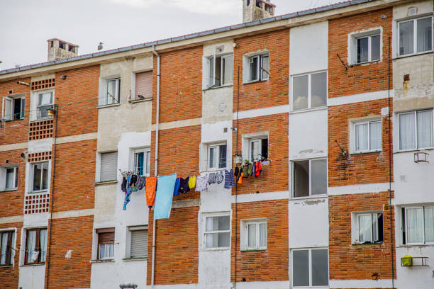 The image showcases a multi-story residential building with a classic, rustic appearance characterized by alternating brick and white concrete walls. The structure is typical of an urban or suburban neighborhood, with multiple windows evenly spaced across the façade. Several balconies are adorned with clotheslines where colorful laundry hangs to dry, adding a vibrant and lived-in feel to the scene. The building's exterior shows signs of age, with some worn areas and peeling paint, reflecting its long-standing presence in the area. There are small details such as electrical wires, street lamps, and the tops of iron fences visible in the foreground. The overall composition captures the essence of everyday life in a residential neighborhood, highlighting the charm and authenticity of community living in an older apartment complex.