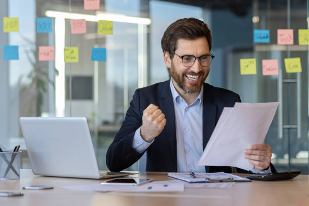 istock Businessman celebrating success while reading document at desk with laptop in modern office setting 2174690080