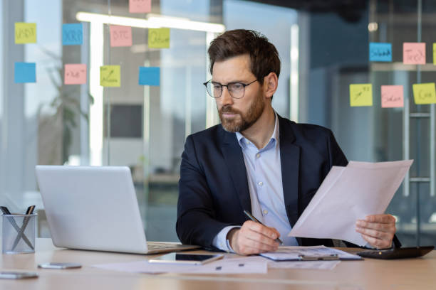 istock Serious mature businessman working in modern office analyzing documents on laptop with focus and determination 2174693578