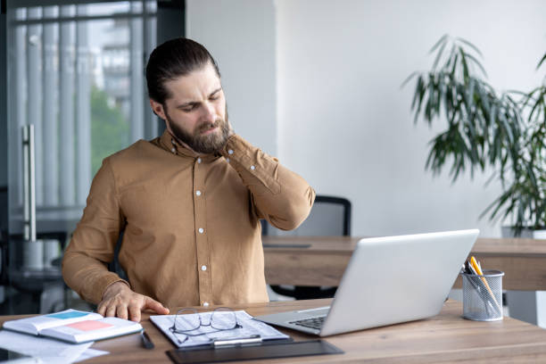 istock Businessman experiencing neck discomfort while working on laptop in modern office environment with documents and glasses on wooden desk. 2174700463