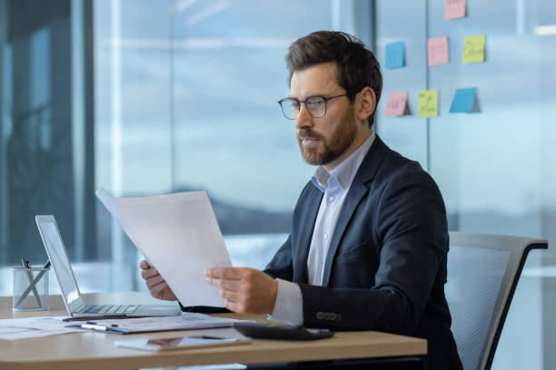 istock Focused businessman in office analyzing documents at desk with computer and smartphone, engaged in important work task 2174710025