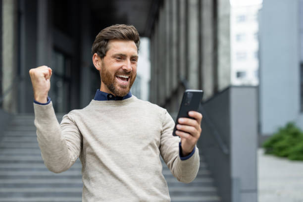 istock Excited man celebrating success while using smartphone in a city setting, expressing happiness and joy with fist pump gesture 2174715578
