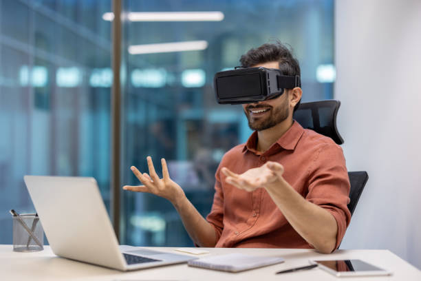 istock Man in office experiencing virtual reality video call using VR headset and laptop with smartphone on desk 2174719623