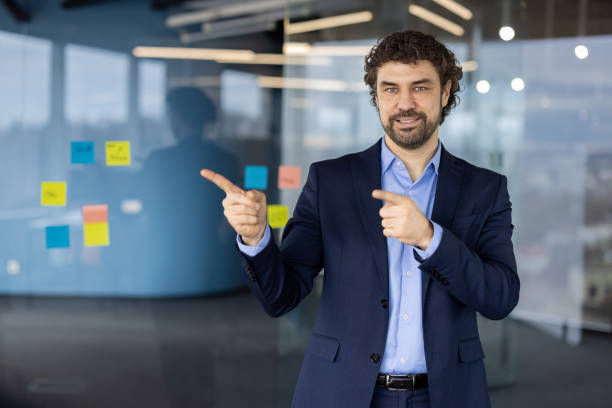 istock Confident businessman in suit points at ideas on office glass wall with sticky notes and smiles at camera 2174722561